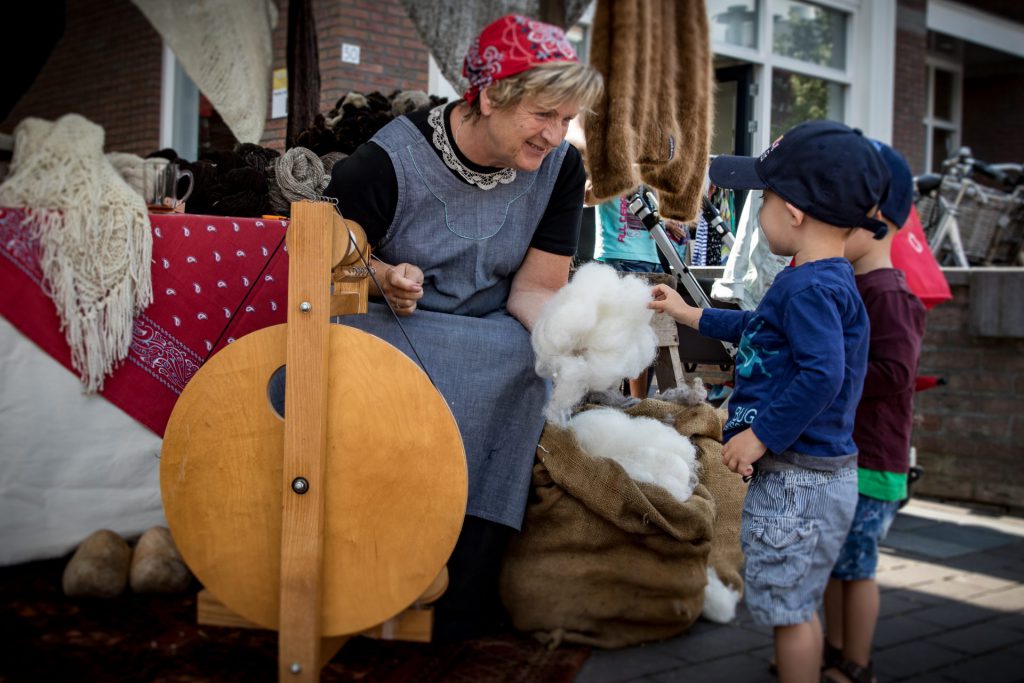 Vrouw demonstreert wolspinnen op een spinnewiel terwijl een kind de wol aanraakt